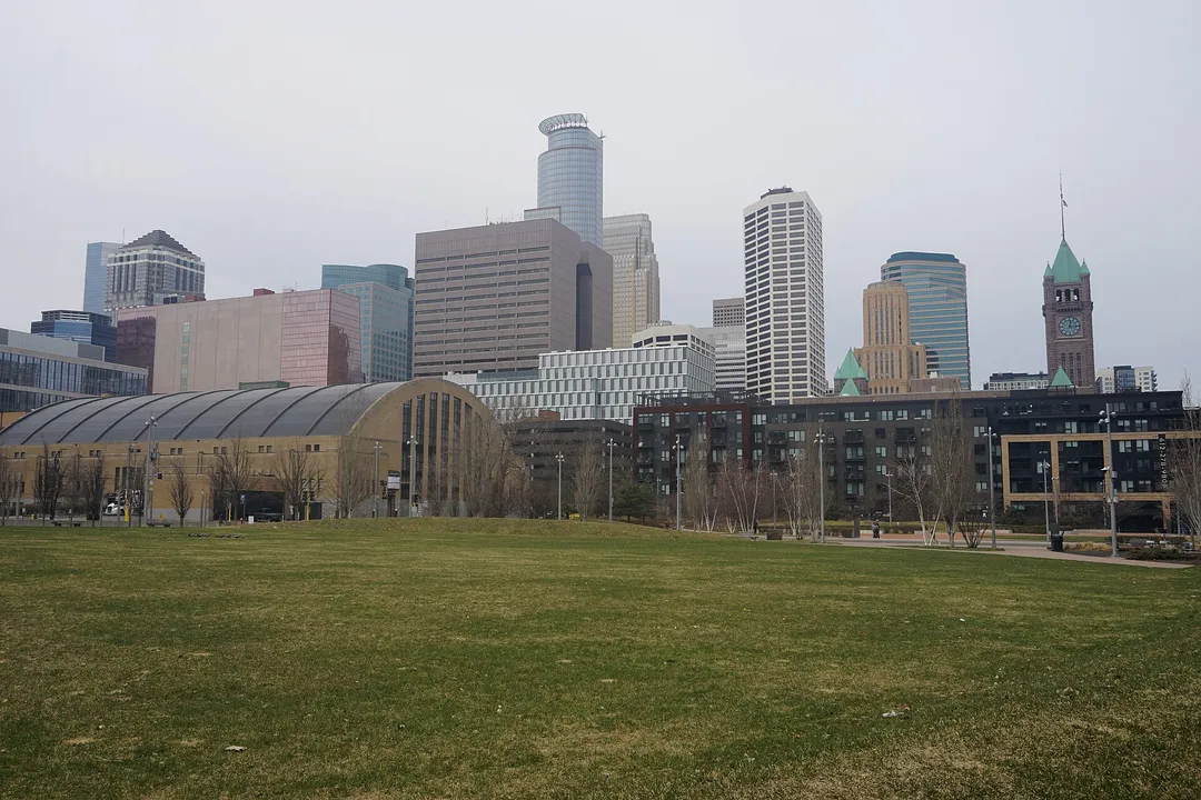 Downtown towers rise behind a glass-roofed building and open lawn in Minnesota.