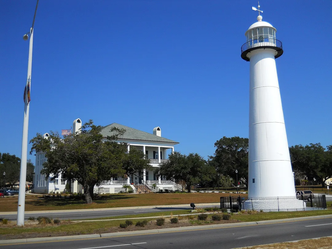 A white lighthouse stands beside a road and a columned house in Mississippi.