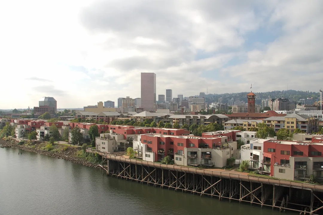 Red-roofed waterfront buildings on pilings sit below Oregon city towers and a wooded hillside.